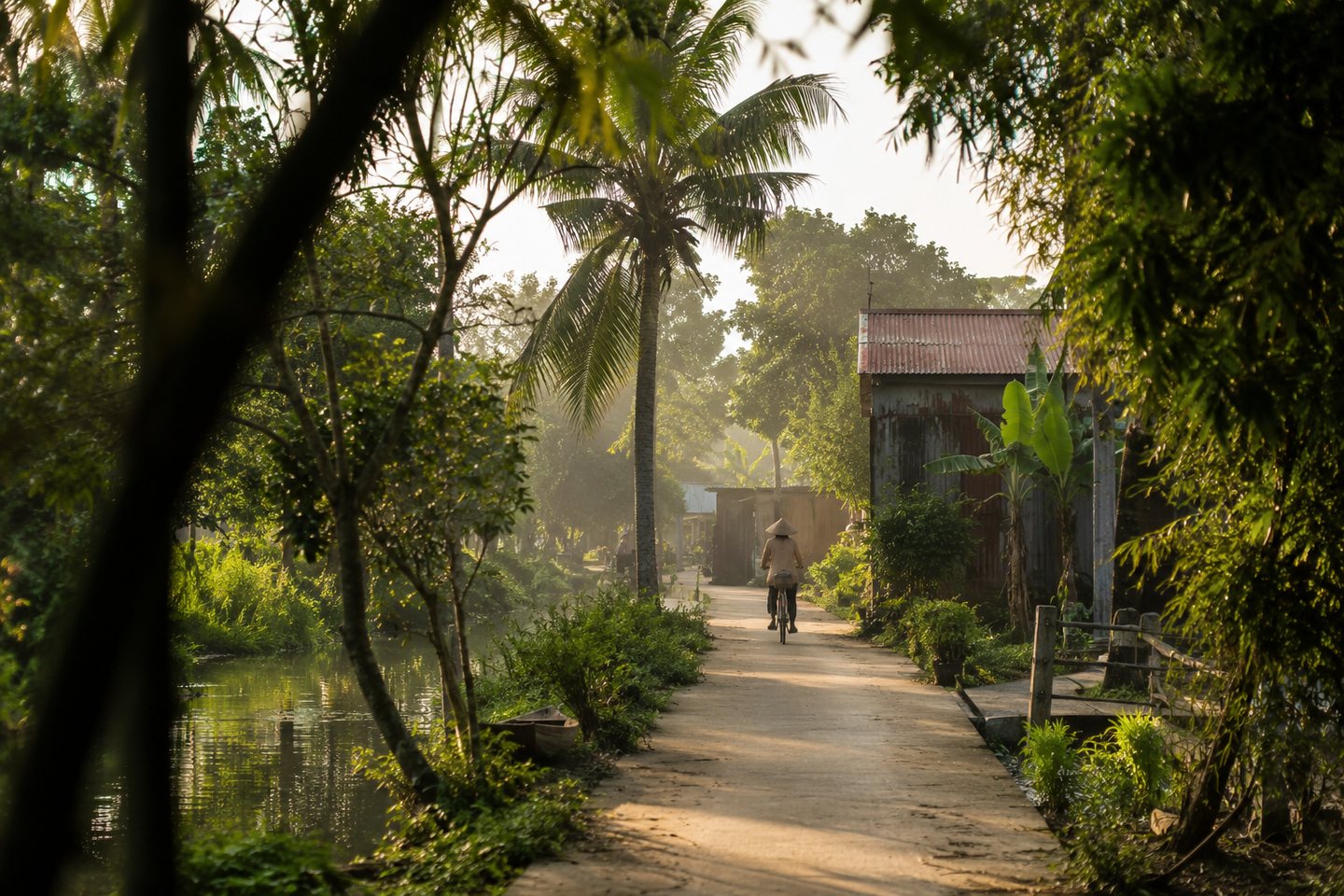 A quiet village lane beside a canal in the Mekong Delta, with bicycle movement in morning light